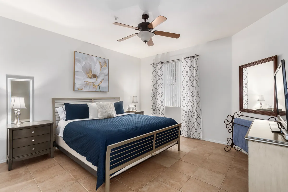 A cozy bedroom with blue bedding, a mirrored dresser, and floral artwork above the bed. Sunlight streams through a curtained window, offering views of the garden, while decorative accents create a welcoming, restful retreat.