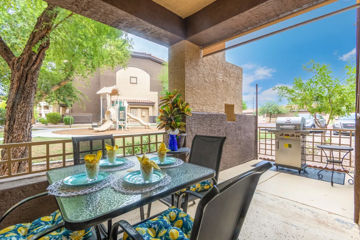 An outdoor patio setup with a dining table ready for a meal, a BBQ grill nearby, and lush greenery all around. In the background, a playground is visible, emphasizing a family-friendly and vibrant outdoor atmosphere. The mood is cheerful and relaxed, suggesting memorable alfresco meals.