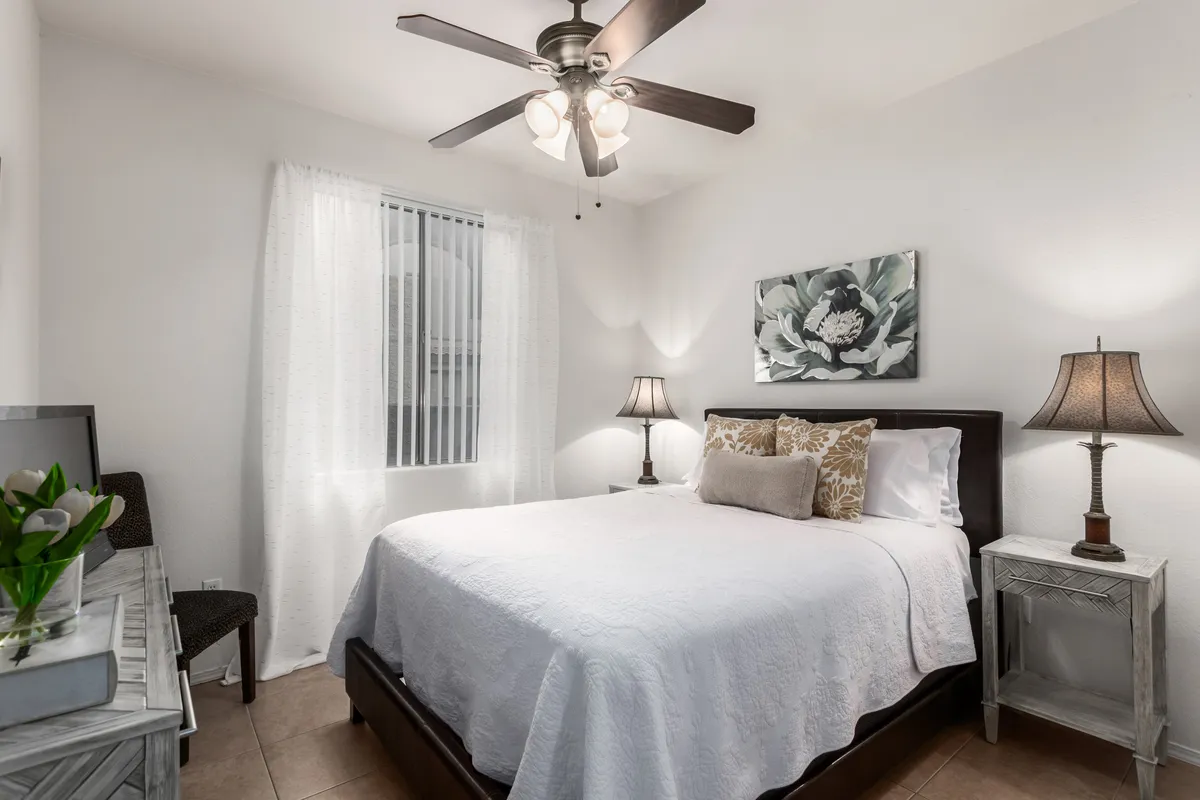 A serene bedroom featuring a neatly made bed with crisp white bedding, decorative pillows, and elegant floral wall art. The bedside tables are topped with lamps, creating a soft, inviting glow perfect for relaxing or reading. The overall ambiance is peaceful and cozy, ideal for restful sleep.