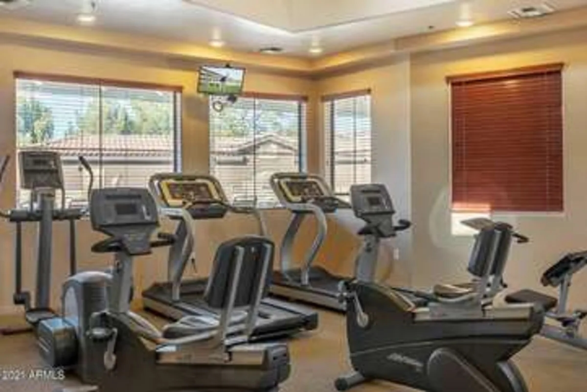 A bright, well-equipped fitness center with several elliptical machines and stationary bikes lined up. Large windows let in natural light and provide a partial view to the outdoors. A television is mounted on the wall for entertainment during workouts. The space feels open, modern, and motivating.