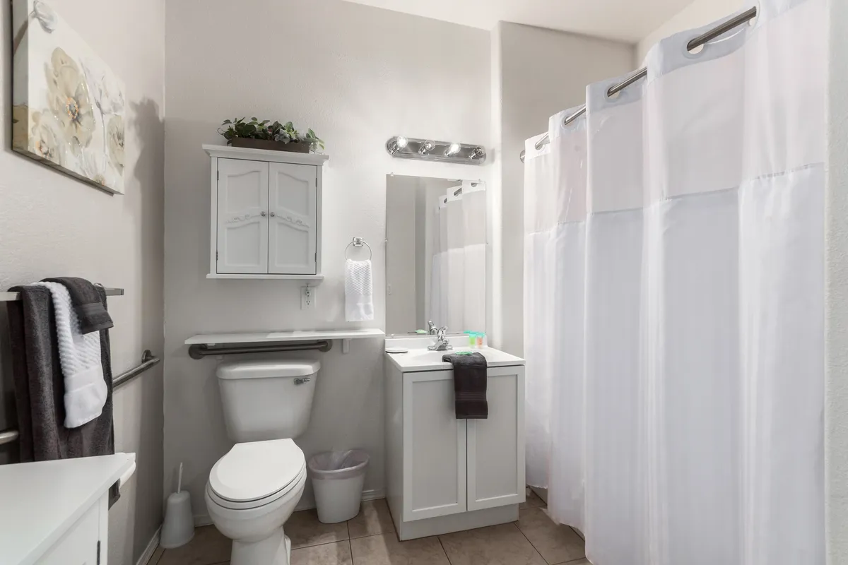 A bright bathroom with a modern vanity and sink, a shower curtain, framed artwork, and a towel rack. The space feels fresh and inviting, perfect for morning routines or a relaxing bath after a long day.