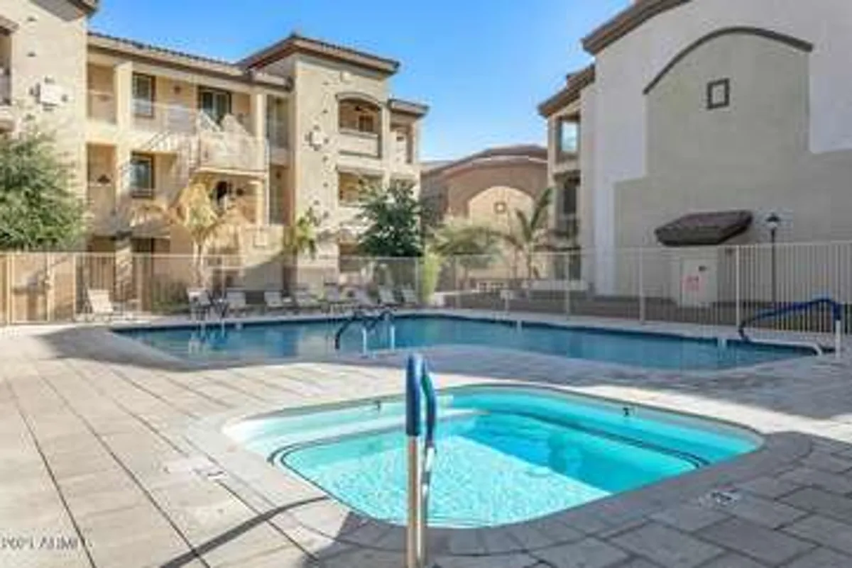A spacious heated swimming pool with an adjoining jacuzzi is surrounded by lush palm trees and the balconies of nearby apartments. People can be seen swimming, sunbathing, and relaxing by the water, contributing to a tranquil, resort-like atmosphere.