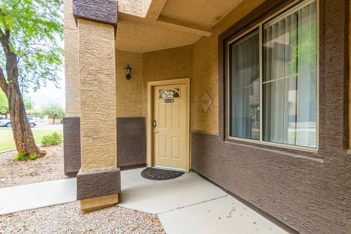 The image shows the inviting front entrance of the condo, featuring a welcoming front door, a large window, textured walls, and thoughtfully arranged landscaping around the entryway. The space conveys a sense of arrival and community, with a porch that hints at moments of outdoor relaxation or friendly greetings with neighbors.